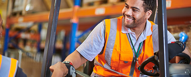 Man on a forklift truck fist bumping a man on the warehouse floor