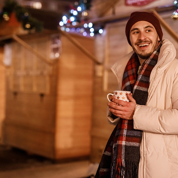 Smiling Man In Long Puffer Jacket Cupping A Mug A Backdrop Of Closed Garlanded Market Sheds And Fairy lights