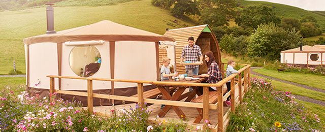 Two Adults And Kids Outside A Yurt In A Field With Wild Fowers