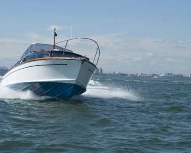 White motorboat at sea sailing towards the camera with spray either side and cityscape in the distance