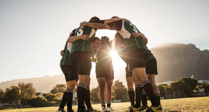 Group of male rugby players in a huddle on the pitch with the sun's rays shining through their arms 